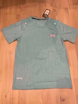 Grön träningströja från Under Armour - Snygg grön träningströja från Under Armour med diskret mönster och färgglad logga på bröstet. T-shirten har rund halsringning och korta ärmar, perfekt för gymmet eller löprundan. Tillverkad i ett lätt och snabbtorkande material.