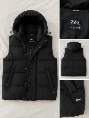 Svart dunväst med huva från ZARA - Snygg svart dunväst från ZARA med avtagbar huva och dragkedja framtill. Västen har två sidofickor och en liten patch med texten 'dmwr.' på ena fickan. Perfekt för lager-på-lager och streetwear-stil.