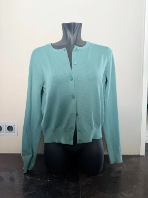 Teal/Ljusgrön basic kofta med rund hals - En stilren teal-färgad kofta med rund halsringning och knäppning framtill. Koftan har långa ärmar och ribbade muddar vid ärmslut och nederkant. Perfekt att slänga över en topp för en enkel och fräsch look.