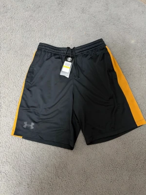 Svarta träningsshorts från Under Armour - Snygga svarta träningsshorts från Under Armour med gula sidopaneler och snörning i midjan. Tillverkade i ett lätt och snabbtorkande syntetmaterial som passar perfekt för sport och aktivitet. Under Armour-logga på vänster ben.