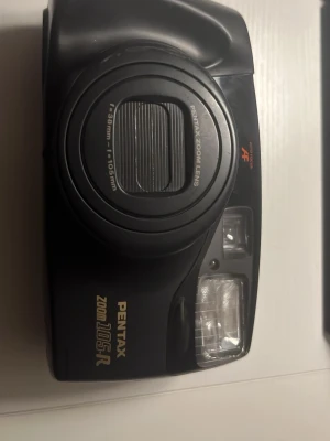Pentax Zoom 105-R kompaktkamera - Pentax Zoom 105-R är en svart kompaktkamera med inbyggd zoomobjektiv (38-105mm) och autofokus. Kameran har en klassisk rektangulär form, inbyggd blixt och flera knappar för olika funktioner på baksidan. Perfekt för dig som gillar analog fotografering.