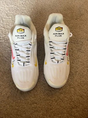 Nike Air Max Plus vita sneakers - Säljer ett par vita Nike Air Max Plus sneakers med svarta och gula detaljer samt en röd swoosh på sidan. Skorna har en sportig design med synlig dämpning i sulan och klassiska Tn-loggan på plösen. Materialet är mesh och syntet för en luftig känsla.