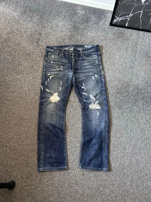 Blå slitna jeans med raka ben - Säljer ett par blå jeans med raka ben och tydliga slitningar. Jeansen har en klassisk femficksmodell och coolt distressed utseende. Perfekta för dig som gillar en avslappnad och trendig stil.