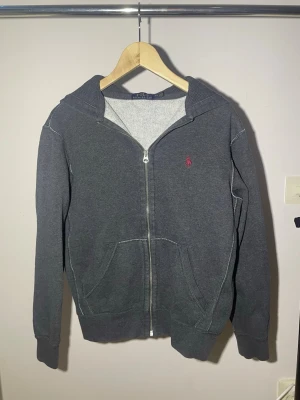 Mörkgrå hoodie från Polo Ralph Lauren - Mörkgrå hoodie med dragkedja från Polo Ralph Lauren. Klassisk design med huva, kängurufickor och den ikoniska röda loggan broderad på bröstet. Tillverkad i mjukt bomullsmaterial som är skönt mot huden. Perfekt för en avslappnad och stilren look.