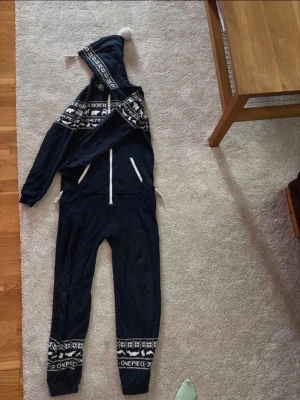 Mörkblå Onepiece jumpsuit med mönster - Mysig mörkblå jumpsuit från Onepiece med vita detaljer och dragkedja framtill. Har huva med tofs och ett vintrigt mönster med björnar och snöflingor över bröst och ben. Denna onepiece passar perfekt för barn 