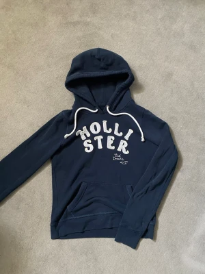 Mörkblå hoodie från Hollister - Snygg mörkblå hoodie från Hollister med broderad logga och texten 'Cali Dreams' på bröstet. Hoodien har en klassisk huva med vita dragsnören och är tillverkad i mjukt bomullsmaterial. Perfekt för en avslappnad och trendig look.