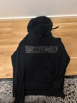 Svart hoodie från BOSS med huva - Snygg svart hoodie från BOSS med huva och dragsko. Framtill finns en bred rand i skinnimitation med BOSS-loggan tryckt över bröstet. Hoodien har känguruficka och ribbade muddar vid ärmslut och nederkant. Perfekt för en avslappnad och trendig look.