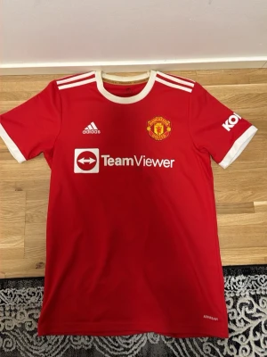 Manchester United Ronaldo tröja - Röd Manchester United t-shirt med vita detaljer, Adidas-logga och TeamViewer-tryck på bröstet. På ryggen står det 'Ronaldo' och nummer 7. Klassisk rund hals och korta ärmar, perfekt för fotbollsfans.
