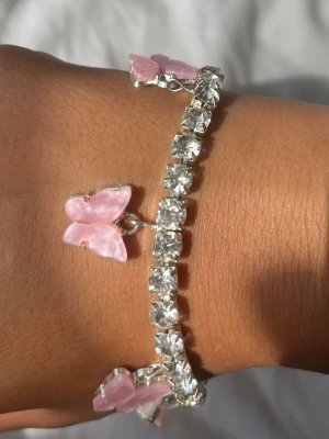 Armband med rosa fjärilar och strass - Glittrigt armband med stora klara strasstenar och fem hängande fjärilar i ljusrosa pärlemor. Kedjan är silverfärgad och fjärilarna ger en söt och trendig look. Perfekt accessoar för att lyfta din outfit med lite extra bling och färg.
