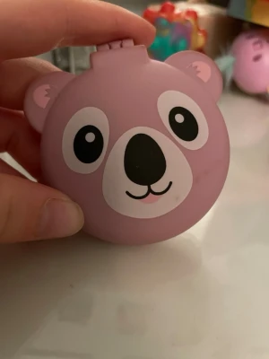 Lila björnformad borste med spegel - Söt och kompakt borste i lila plast med björndesign. Inuti finns en spegel och en utfällbar borste med rosa och svarta piggar. Perfekt att ha i väskan för snabba fixar. Praktisk och lekfull accessoar för hårvård.