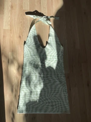 Grönvit kort klänning med knyt - Säljer en kort klänning i ljusgrönt och vitt med grafiskt mönster. Klänningen har djup v-ringning och knyt i nacken, vilket ger en snygg halterneck-look. Perfekt för varma dagar och sommarens alla tillfällen.