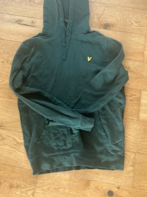 Mörkgrön hoodie från Lyle & Scott - Snygg mörkgrön hoodie från Lyle & Scott med klassisk gul logga på bröstet. Hoodien har huva med dragsko och en stor magficka. Tillverkad i mjukt bomullsmaterial som är skönt att bära. Perfekt för en avslappnad och stilren look.