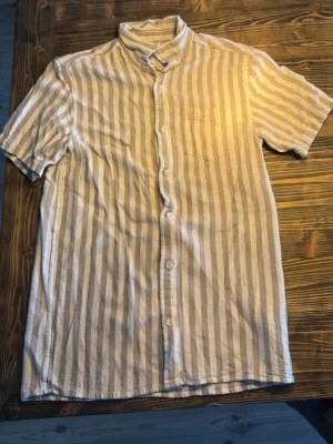 Randig kortärmad skjorta beige/vit - Kortärmad skjorta med vertikala beige och vita ränder. Skjortan har klassisk krage, bröstficka och knäppning framtill. Tillverkad i ett lätt och luftigt material, perfekt för varmare dagar.