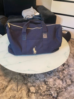 Mörkblå weekendbag från Polo Ralph Lauren - Snygg mörkblå weekendbag i canvas från Polo Ralph Lauren med klassisk gul logga framtill. Väskan har dubbla handtag och dragkedja upptill. Perfekt storlek för kortare resor eller gymmet. Enkel och stilren design som passar till det mesta.