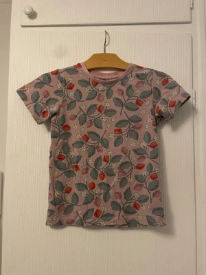Blommig t-shirt från Polarn O. Pyret - Söt t-shirt från Polarn O. Pyret i storlek 122 med blommigt och jordgubbsmönster på ljusrosa botten. T-shirten har korta ärmar och rund halsringning. Perfekt för dig som gillar färgglada och lekfulla plagg.