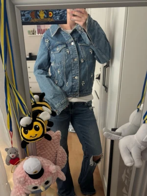 Blå jeansjacka med pärldetaljer - Säljer en snygg blå jeansjacka med pärldetaljer på framsidan och axlarna. Klassisk modell med krage, bröstfickor och silverfärgade knappar. Jackan har en avslappnad passform och är perfekt för dig som vill sticka ut lite extra.