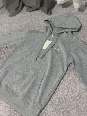 Zara - Snygg grå hoodie med huva och dragsnören. Har en broderad logga på bröstet och en stor magficka framtill. Tillverkad i mjukt sweatshirtmaterial som känns skönt mot huden. Perfekt för en chill och avslappnad stil.