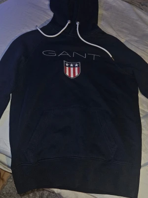 Mörkblå hoodie från GANT - Snygg mörkblå hoodie från GANT med vit dragsko i huvan och stor ficka framtill. På bröstet finns GANTs logga. Inga deffekter finns på den. Storlek XS