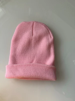 Ljusrosa ribbad mössa - helt NY - Helt NY stilren ljusrosa mössa i ribbstickad bomullsmix med uppvikt kant. Perfekt för att addera lite färg till din outfit och hålla värmen under kyliga dagar. Enkel och klassisk design som passar till det mesta. Helt NY, oanvänd. 