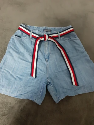 Blå jeansshorts Tommy Hilfiger - Snygga ljusblå jeansshorts från Tommy Hilfiger med ett randigt bälte i rött, vitt och blått. Klassisk femficksmodell och avslappnad passform, perfekta för varma dagar. Bältet ger en extra färgklick och cool detalj till outfiten.