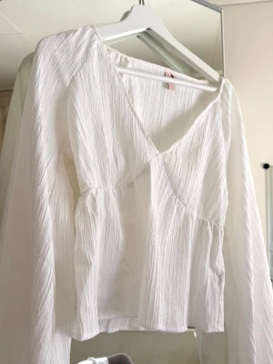 nelly blus  - Säljer en vit blus med v-ringning och lång ärm. Blusen har ett lätt rynkat tyg med struktur och en avslappnad passform. Perfekt att styla med jeans eller kjol för en trendig look. tryck på köp nu 💕