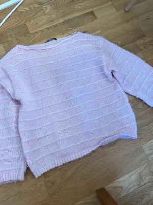 Ljusrosa stickad tröja från Bik Bok - Söt ljusrosa stickad tröja från Bik Bok med tunna vita ränder. Tröjan har en avslappnad passform, rund halsringning och ribbade muddar vid ärmslut och nederkant. Perfekt för lager på lager och ger en mjuk, fluffig känsla.