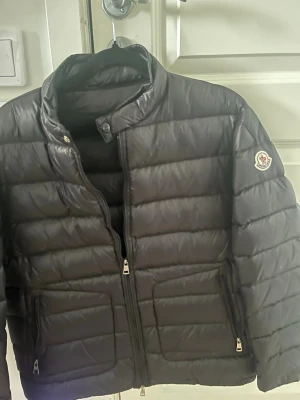 Svart dunjacka från Moncler - Snygg svart dunjacka från Moncler med quiltad design och hög krage. Jackan har dragkedja framtill, två sidofickor med dragkedja och Moncler-logga på ärmen. Perfekt för kalla vinterdagar och riktigt stilren look.