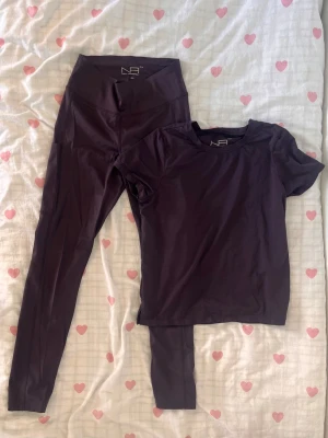 Mörklila träningsset från New Apparel - Säljer ett mörklila träningsset från New Apparel i storlek XS. Setet består av ett par höga tights och en matchande kortärmad t-shirt i stretchigt funktionsmaterial. Perfekt för gymmet eller löpning. Snygg, enkel design med diskret struktur i tyget.