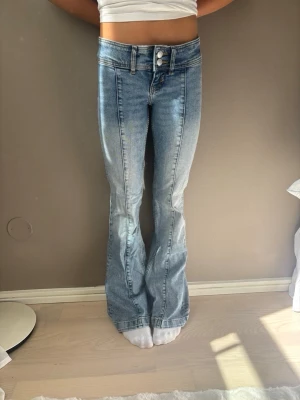 Ljusblå bootcut jeans med hög midja - Säljer ett par ljusblå bootcut jeans med hög midja och dubbla knappar framtill. Jeansen har klassiska fickor både fram och bak samt en snygg tvättad look. Perfekta för dig som gillar en retro och avslappnad stil.