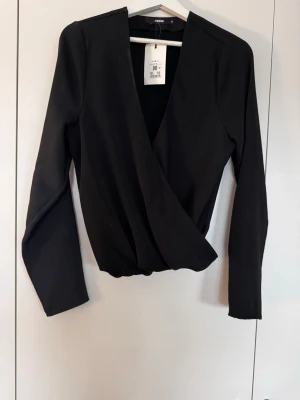 Svart omlottopp med lång ärm - Stilren svart omlottopp med djup v-ringning och långa ärmar. Toppen har en elegant omlottdesign som ger en snygg siluett och passar perfekt till många olika looks. Tillverkad i ett mjukt och följsamt material.