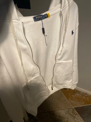 Vit hoodie från Polo Ralph Lauren - Säljer en vit hoodie från Polo Ralph Lauren med dragkedja, huva med snören och känguruficka. Klassisk blå logga broderad på bröstet. Tillverkad i mjukt material som känns skönt mot huden. Perfekt för en avslappnad och stilren look.