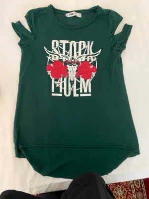 Grön t-shirt med tryck och slitsade ärmar - Cool grön t-shirt med vitt tryck där det står 'Stockholm' och motiv av horn, rosor och dödskalle. T-shirten har korta, slitsade ärmar och rund hals. Materialet känns mjukt och skönt, perfekt för en avslappnad stil.