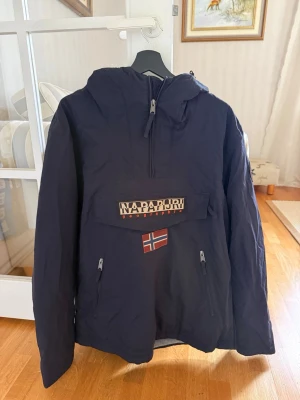 Mörkblå vindjacka från Napapijri - Snygg mörkblå vindjacka från Napapijri med stor ficka framtill,  Jackan har huva och är tillverkad i slitstarkt material, perfekt för blåsiga dagar. Regular fit och fleecefoder på insidan för extra komfort. Passar bra till skidresor! 