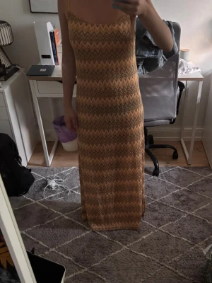 Lång zigzag-mönstrad klänning - Säljer en lång klänning med smala axelband och zigzag-mönster i beige, gult och brunt. Klänningen har en rak passform och är perfekt för varma dagar. Materialet känns lätt och luftigt, vilket gör den skön att bära. Storleken är L men för mig som är 165 och väger ca 58 kg sitter den som M