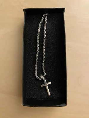 Ropechain halsband med korshänge 60cm - Snyggt halsband med tvinnad kedja och ett stilrent korshänge. Kedjan har en modern look och passar perfekt för dig som gillar minimalistiska accessoarer. Materialet ser ut att vara metall och designen är enkel men ändå trendig.