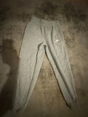 Gråa mjukisbyxor från Nike - Säljer ett par klassiska gråa mjukisbyxor från Nike med resår i midjan och muddar vid bensluten. Byxorna har en enkel och clean look med Nike-logga på vänster lår. Perfekta för chill dagar eller träning.