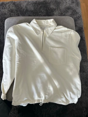 Vit henley med dragkedja från Samsøe Samsøe - Säljer en stilren vit henley från Samsøe Samsøe med dragkedja framtill, klassisk krage och bröstficka. Skjortan har långa ärmar och dragsko i nederkant för en avslappnad look. Perfekt till jeans eller chinos.