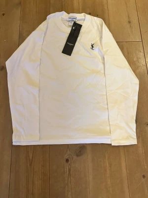 Vit långärmad topp från Saint Laurent - Vit långärmad topp från Saint Laurent med rund halsringning och svart broderad YSL-logga på bröstet. Tillverkad i mjuk bomull för en bekväm känsla. Perfekt för dig som gillar stilrena och klassiska plagg. Helt oanvänd med inga defekter. Fraktar inom 1-2 dagar!