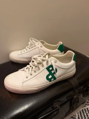 Vita sneakers med grön logga - Snygga vita sneakers från BOSS med grön logga på sidan och grön detalj vid hälen. Skorna har klassisk låg profil, vita snören och ovandel i slätt läder. Perfekta för en clean och stilren look.