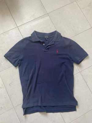 Marinblå pikétröja från Polo Ralph Lauren - Klassisk marinblå pikétröja från Polo Ralph Lauren med korta ärmar och krage. Tröjan har två knappar vid halsen och den ikoniska röda logotypen broderad på bröstet. Perfekt till jeans eller shorts för en clean look. Tröjan är använd men fortfarande i fint skick. Vet inte riktigt storlek men passar mig som brukar bära S