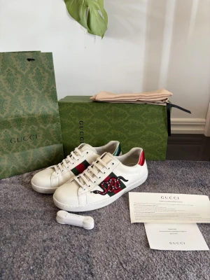 Gucci Ace vita sneakers med orm - Säljer ett par vita Gucci sneakers i läder med ikonisk röd och svart orm broderad på sidan. Skorna har använts ett få antal gånger. Allt på bilden med följer