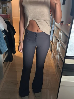 Mörkgrå bootcut byxor med knappar - Säljer ett par mörkgrå bootcut byxor med hög midja och snygga knappar framtill. Byxorna har fickor bak med knappdetalj och är tillverkade i ett stretchigt material som sitter riktigt snyggt på kroppen.