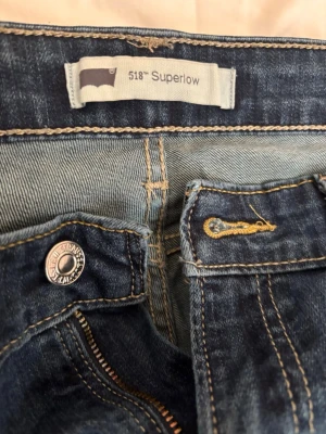 Levi's 518 Superlow blå jeans - Ett par klassiska Levi's 518 Superlow jeans i blå denim med låg midja och raka ben. Jeansen har tydliga kontrastsömmar, fem fickor och Levi's ikoniska bakfickor med bågformad söm. Perfekta för en avslappnad och trendig look.