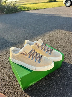 Sjukt fina Beige sneakers från Diemme i mocka - Snygga beige sneakers från Diemme i mjuk mocka med vita sulor och bruna vandringsinspirerade snören. Skorna har silverfärgade metalldetaljer vid snörningen och en stilren, låg profil. Perfekta för dig som gillar en clean och trendig look.