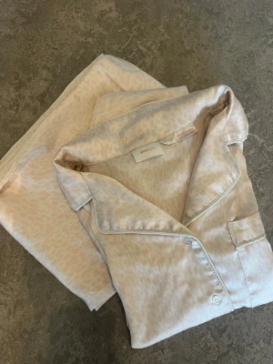 Ljusbeige pyjamas med leopardmönster - Säljer en lyxig ljusbeige pyjamas med subtilt leopardmönster. Setet består av en skjorta med krage, knappstängning och bröstficka samt matchande byxor med resår i midjan. Materialet är glansigt och mjukt, perfekt för mysiga kvällar hemma.