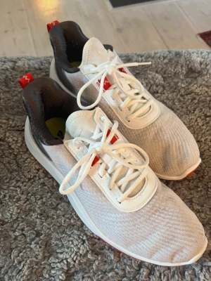 Vita golfskor med röd sula - Säljer ett par vita sneakers med mesh-ovandel, röda detaljer och röd yttersula. Skorna har vita snören, svart vadderad häl och är lätta samt ventilerande. Perfekta för dig som gillar sportig stil och komfort. Endast använda en gång och tvättade efter det.