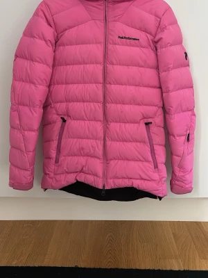 Rosa skidjacka från Peak Performance - Säljer en rosa dunjacka från Peak Performance med huva och två dragkedjeförsedda fickor framtill. Jackan har quiltad design och logga på bröstet. Perfekt för kalla vinterdagar och riktigt skön att bära.
