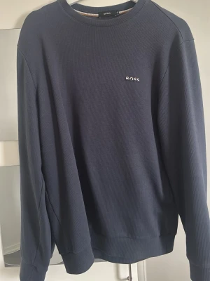 Mörkblå sweatshirt från BOSS - Snygg mörkblå sweatshirt från BOSS med diskret logga broderad på bröstet. Tröjan har rund halsringning, långa ärmar och ribbade muddar vid ärmslut och nederkant. Perfekt för en clean och stilren look.