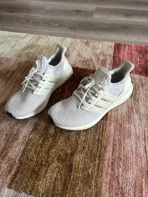 Vita Adidas Ultraboost sneakers - Snygga vita Adidas Ultraboost sneakers med stickad ovandel och klassiska tre ränder på sidan. Skorna har vita snören och en bekväm, responsiv sula. Yttersulan är svart med Continental-mönster för extra grepp. Perfekta för dig som gillar stilrena och sportiga sneakers. Passar storlek 43,5, 44