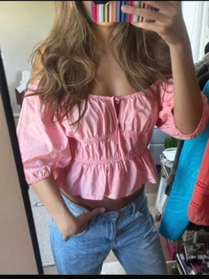 Ljusrosa croppad blus med volang - Supersöt ljusrosa blus med puffiga korta ärmar, volangkant och smock i midjan. Toppen har en liten knytning framtill och är croppad vilket ger en trendig look. Perfekt för dig som vill ha en feminin och somrig stil.